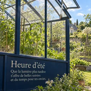 Une lucarne automatique s’ouvre dans une serre verdoyante à la lumière du printemps, avec le texte « Heure d’été Que la lumière plus tardive t’offre de belles soirées et du temps pour tes envies. ».